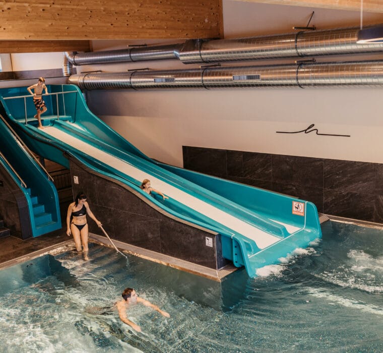 Die Eltern schwimmen im Indoorpool während die Kinder die Trio-Slide Rutsche runter rutschen.