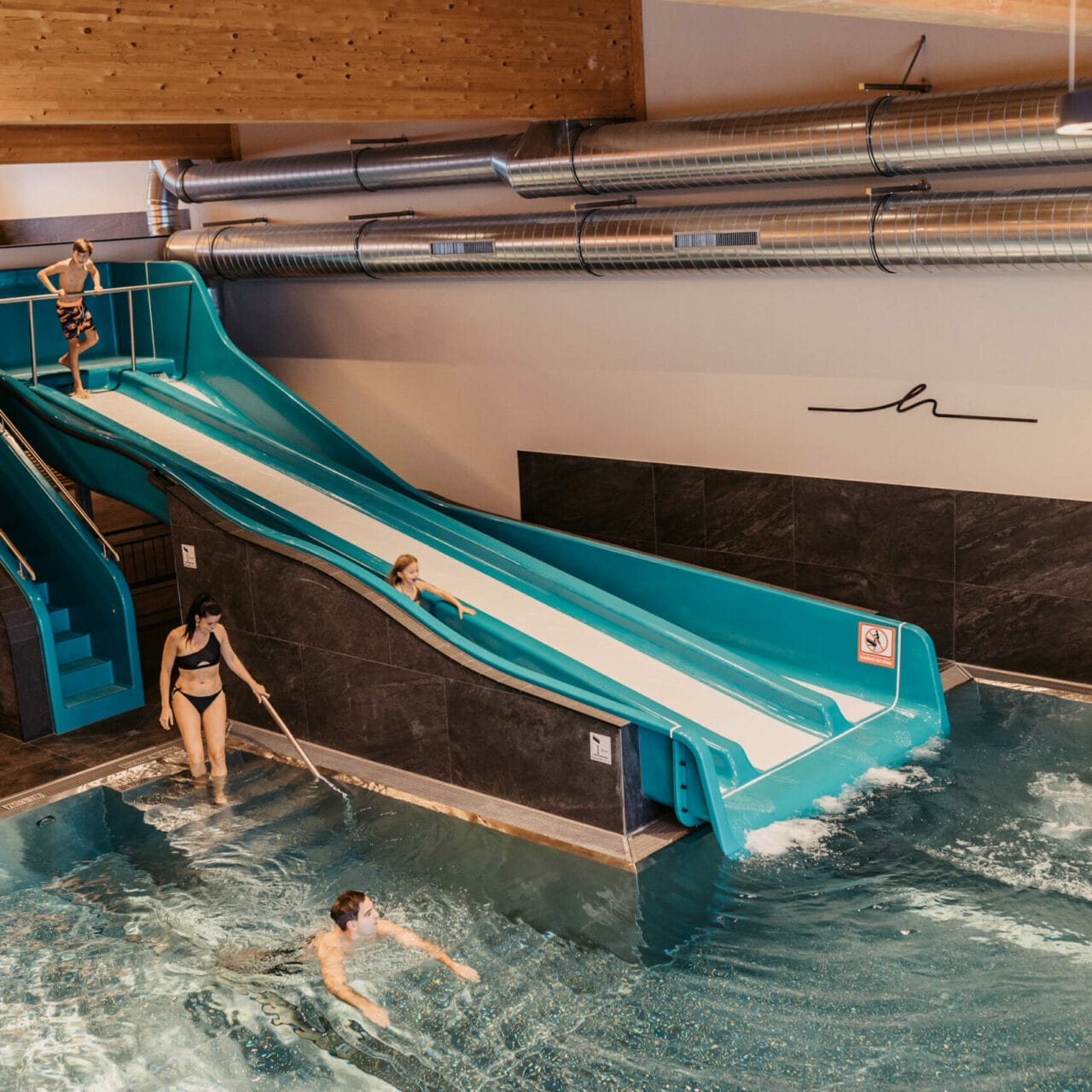 Die Eltern schwimmen im Indoorpool während die Kinder die Trio-Slide Rutsche runter rutschen.