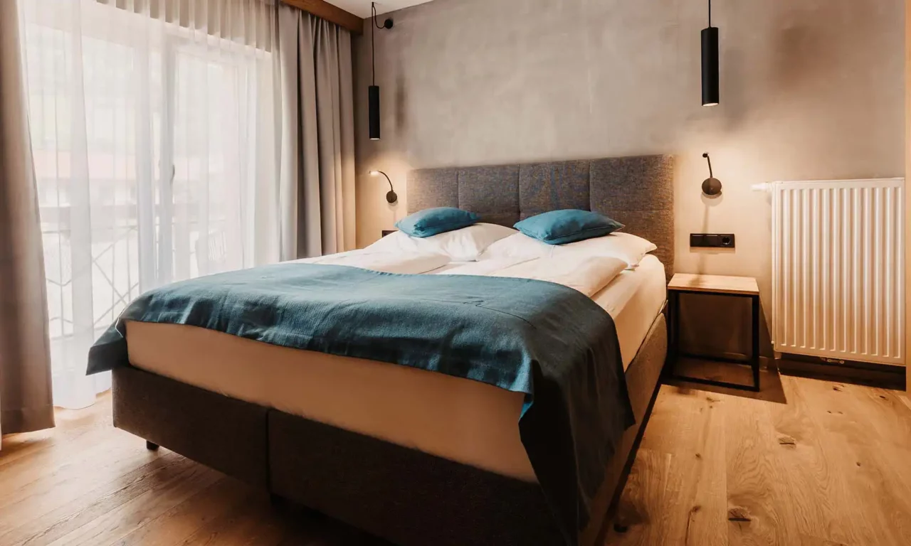 Modern eingerichtetes Schlafzimmer mit gemütlichem Boxspringbett, stilvollen Nachttischen, minimalistischen Hängelampen und Tageslicht durch bodentiefe Fenster mit Vorhängen – alpiner Wohnkomfort im Designstil.
