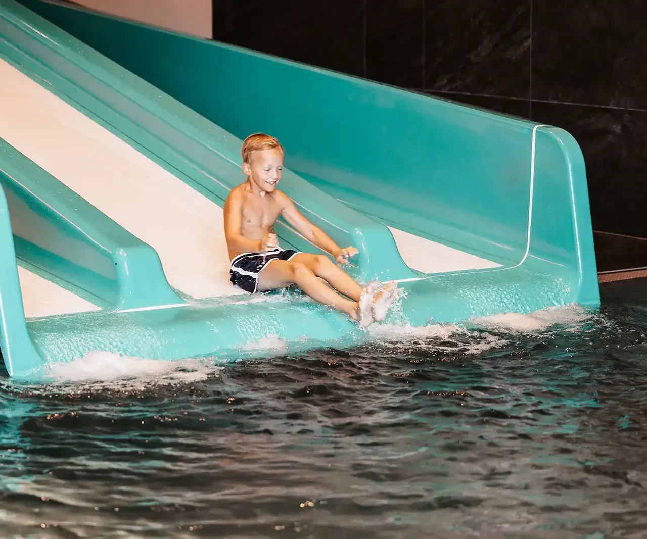 Fröhlicher Junge rutscht lachend eine türkisfarbene Wasserrutsche in ein beheiztes Hallenbad hinunter – Indoor-Wasserspaß für Kinder im Familienurlaub.