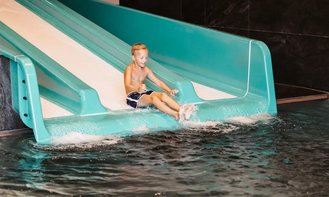 Fröhlicher Junge rutscht lachend eine türkisfarbene Wasserrutsche in ein beheiztes Hallenbad hinunter – Indoor-Wasserspaß für Kinder im Familienurlaub.