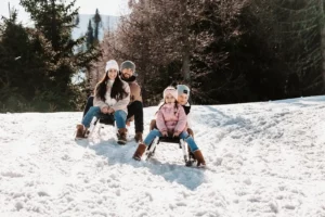 Familie beim Rodeln auf einem verschneiten Hang, lachend und in Winterkleidung.