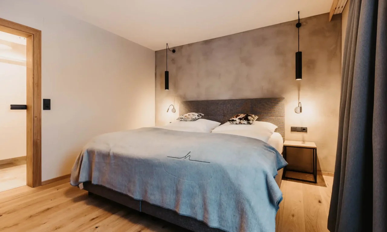 Hyggeligt soveværelse med moderne boxspring-seng, minimalistisk belysning, trægulv og bløde naturfarver - stilfuldt design med alpin flair.