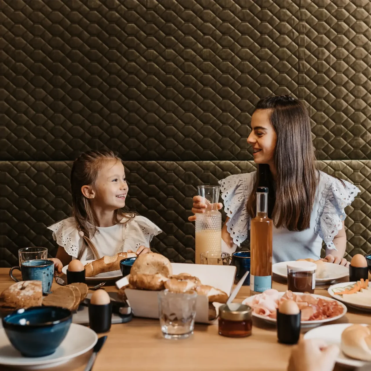 Fröhliche Familie beim gemeinsamen Frühstück – zwei Mädchen lachen und genießen frische Brötchen, Eier und Saft in gemütlicher Atmosphäre.