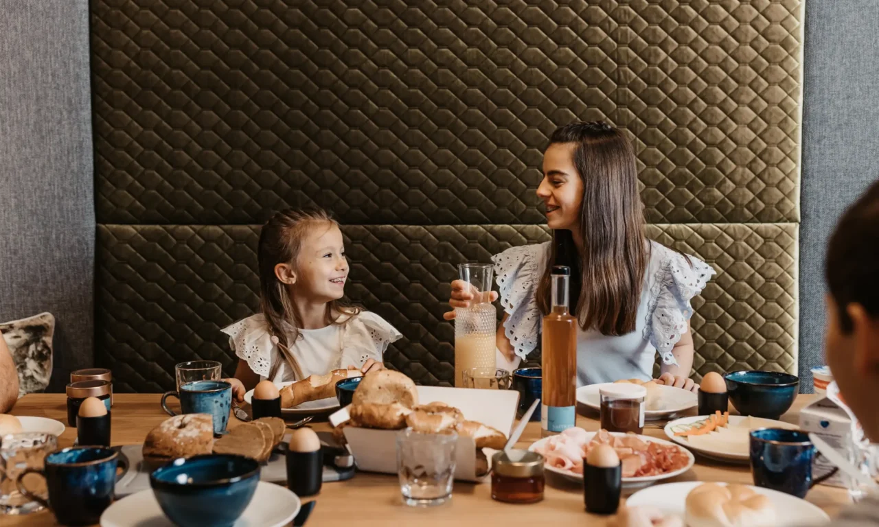 A cheerful family enjoying breakfast together – two girls laugh and enjoy fresh bread rolls, eggs, and juice in a cozy atmosphere.