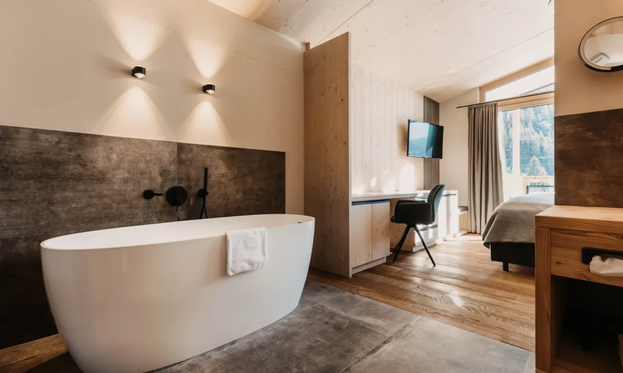 Modern hotel room with freestanding bathtub, stylish wood design, desk, and panoramic windows overlooking the snow-covered mountain landscape.