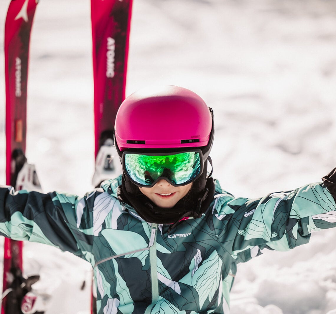 Lächelndes Kind mit pinkem Helm und Atomic-Ski sitzt im Schnee und genießt den Wintertag.