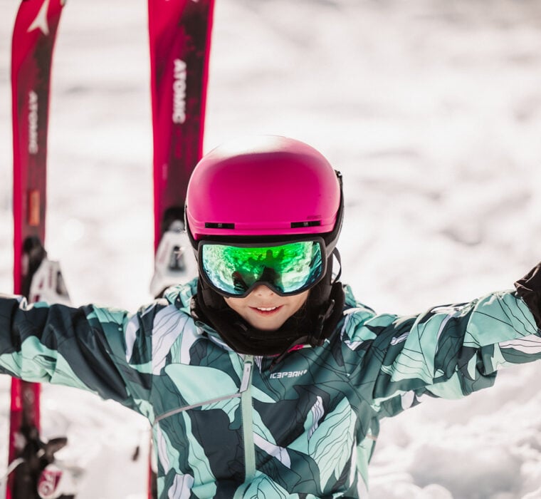 A smiling child wearing a pink helmet and Atomic skis sits in the snow and enjoys the winter day.