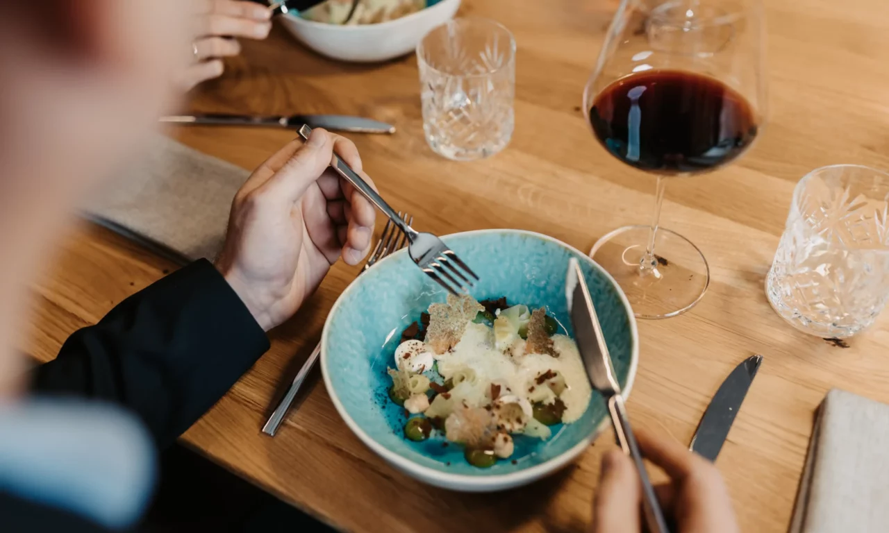 Gourmetgerecht in een turquoise schaal op een houten tafel, stijlvol opgemaakt met schuim, groenten en knapperige elementen; op de voorgrond een persoon die aan het eten is, daarnaast een glas rode wijn en waterglazen.