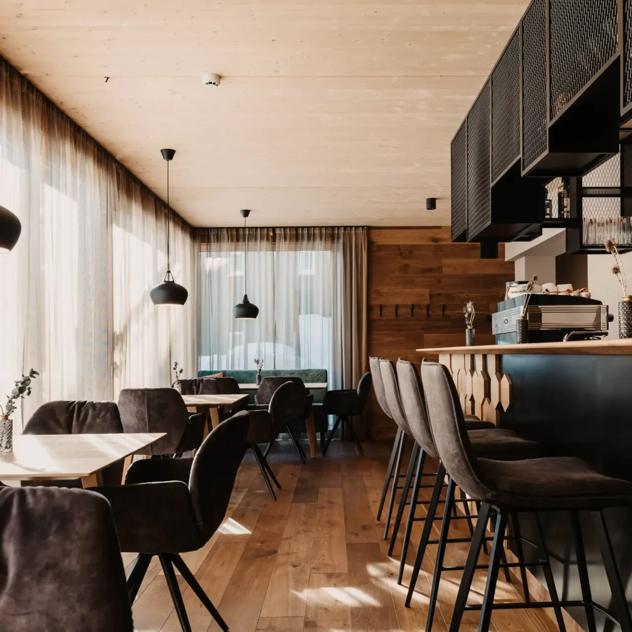 Moderní interiér restaurace s útulnými sametovými židlemi, světlým dřevěným obložením, elegantním barovým pultem a velkými okny s průsvitnými závěsy - stylová kombinace alpské elegance a městského designu.