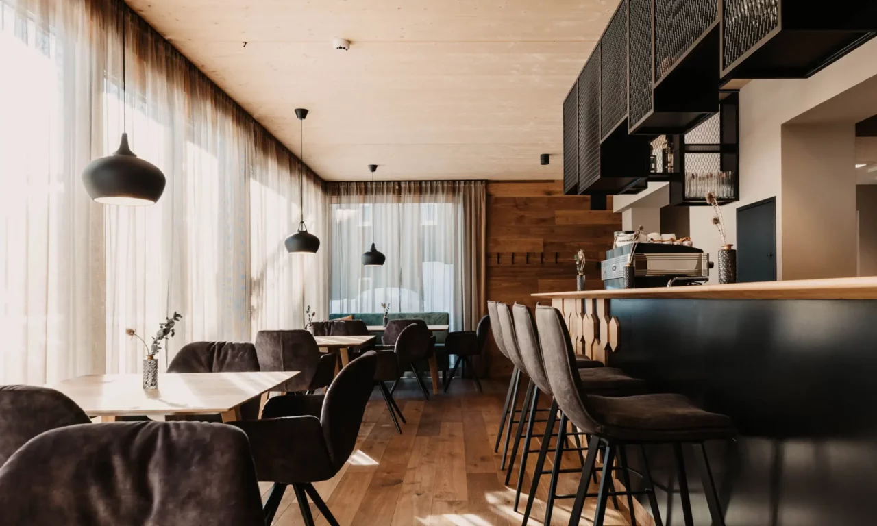 Modern restaurantinterieur met gezellige fluwelen stoelen, lichte houten lambrisering, elegant bargedeelte en grote ramen met doorschijnende gordijnen - een stijlvolle mix van alpine chic en stedelijk design.