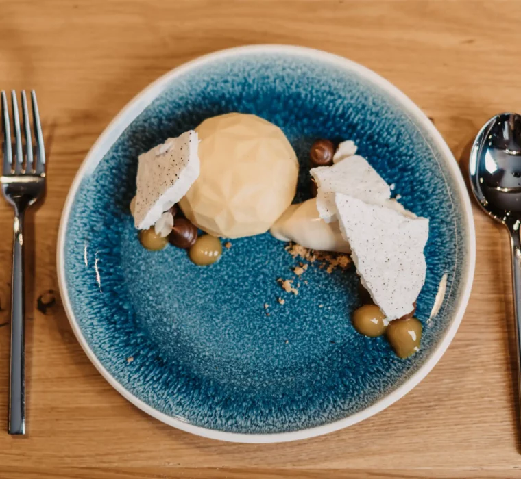 Modern dessert served on a blue ceramic plate with geometric praline, crispy meringue garnish, hazelnuts, cream and fruit gel, served on a light-coloured wooden table.