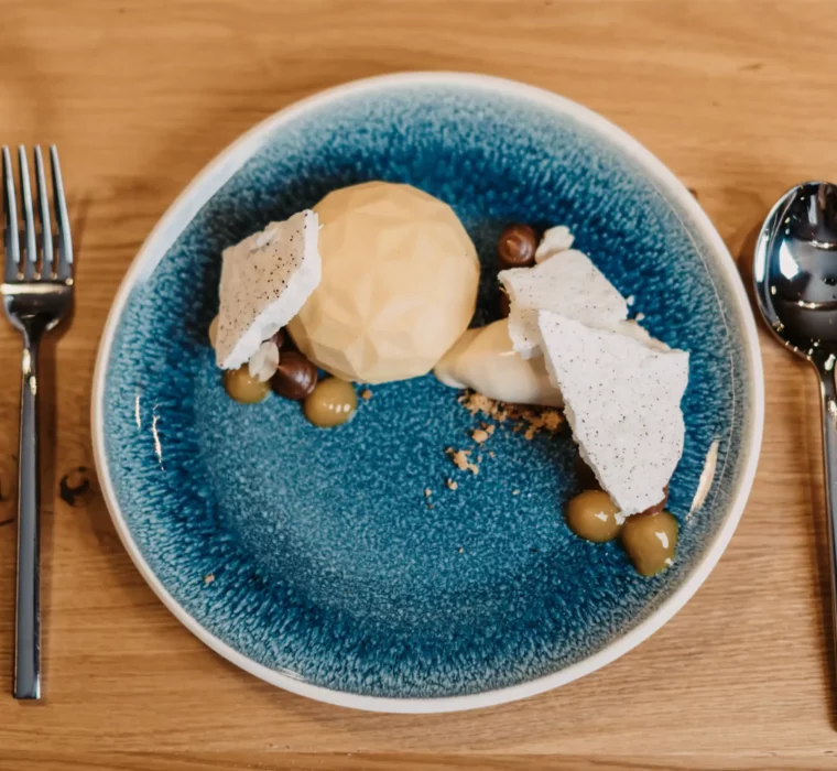 Modern angerichtetes Dessert auf einem blauen Keramikteller mit geometrischer Praline, knuspriger Baiser-Garnitur, Haselnüssen, Creme und Fruchtgel, serviert auf hellem Holztisch.