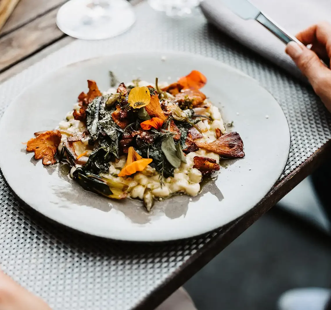 Fein angerichtetes Risotto mit Pfifferlingen, frischen Kräutern und essbaren Blüten auf einem hellen Steingutteller, serviert auf einem stilvoll gedeckten Holztisch.