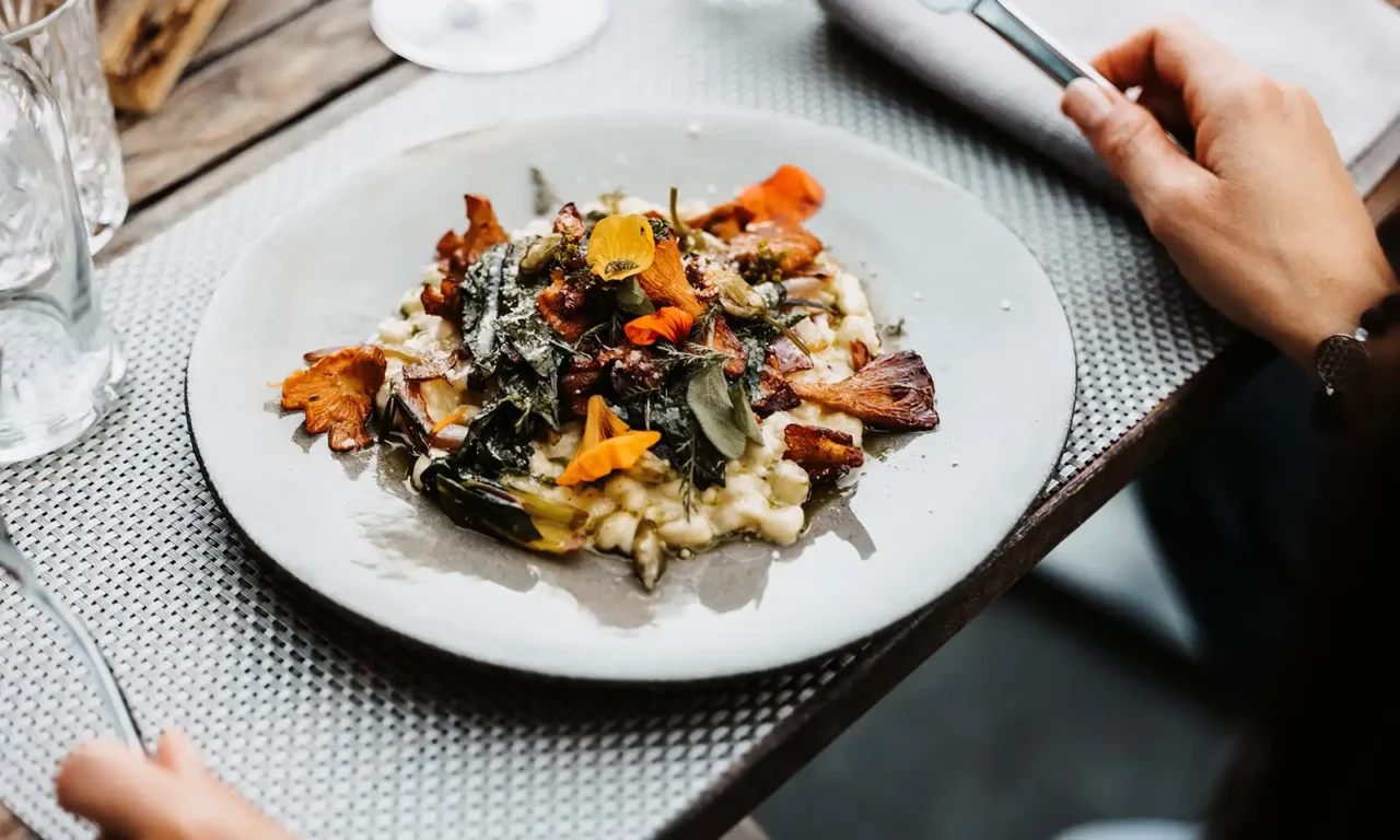 Fijn bereide risotto met cantharellen, verse kruiden en eetbare bloemen op een lichtgekleurd aardewerken bord, geserveerd op een stijlvol gedekte houten tafel.