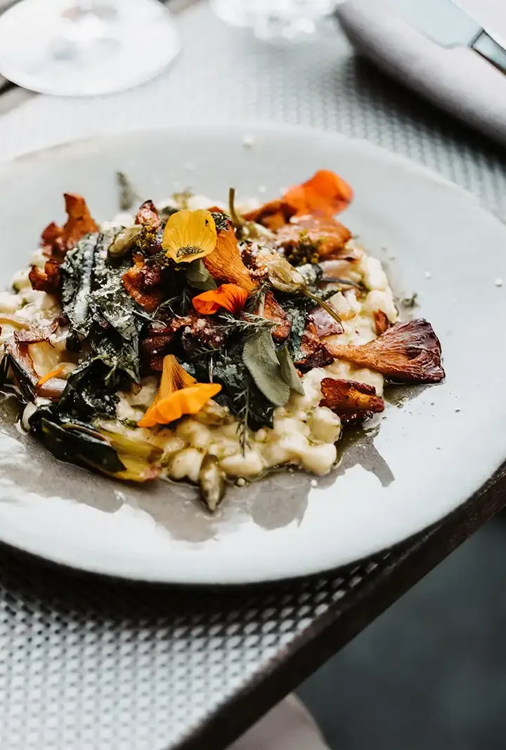 Fein angerichtetes Risotto mit Pfifferlingen, frischen Kräutern und essbaren Blüten auf einem hellen Steingutteller, serviert auf einem stilvoll gedeckten Holztisch.