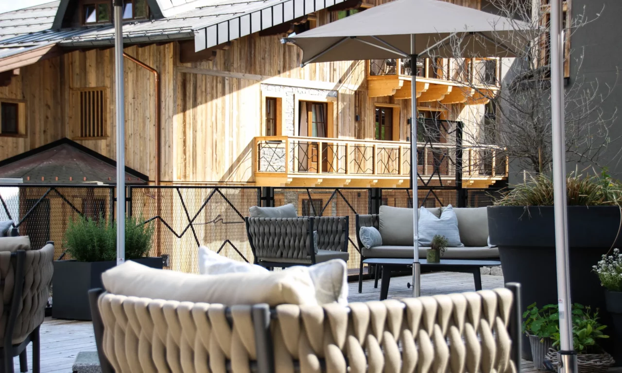 Stijlvol ingericht zonneterras met gezellig gestoffeerd meubilair, parasols en groene plantenbakken voor de gevel van een alpenchalet van natuurlijk hout.