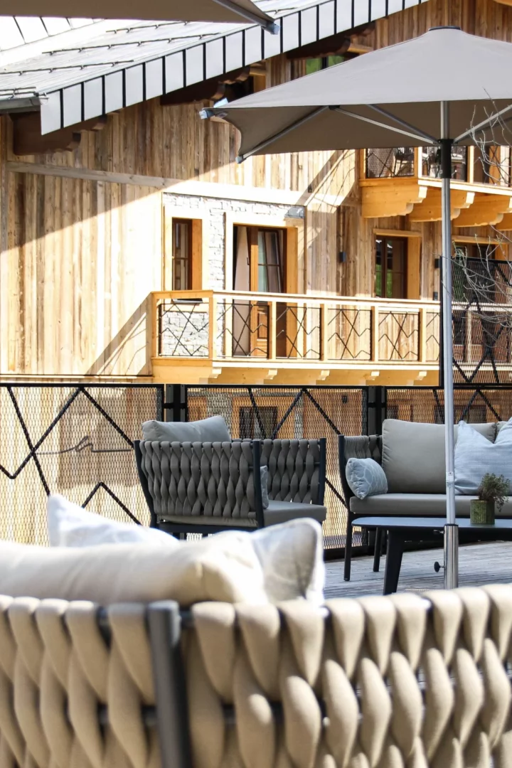 Stilvoll möblierte Sonnenterrasse mit gemütlichen Polstermöbeln, Sonnenschirmen und grünen Pflanzgefäßen vor der Fassade eines alpinen Chalets aus Naturholz.