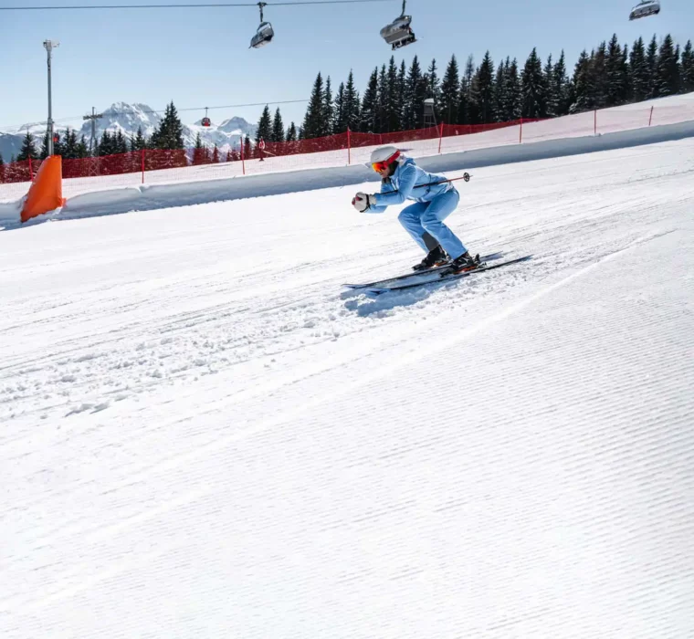 Skiër in lichtblauwe skikleding skiet in een sportieve houding over een vers geprepareerde piste. Besneeuwde bomen, stoeltjesliften en een bergpanorama op de achtergrond.