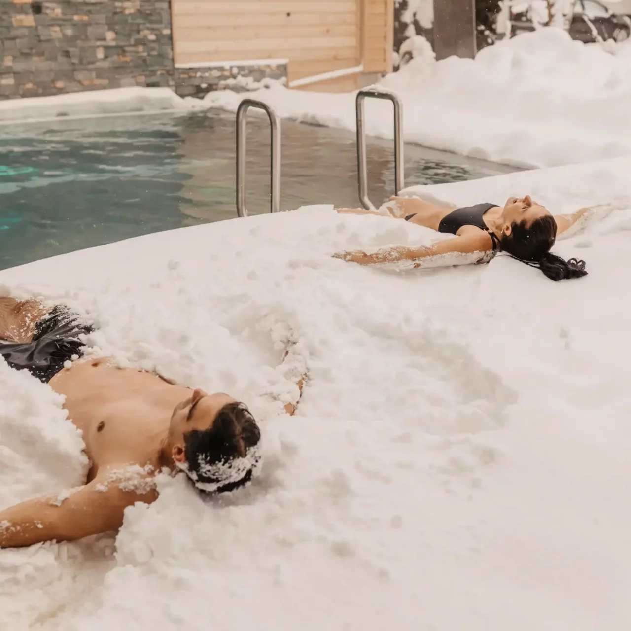 Hofgut-wagrain-apartment-Hallenbadaußenbecken-schnee-302 Two adults in swimwear lie relaxed in the snow next to a heated outdoor pool. The snow envelops their bodies while warm clouds of steam rise from the water in the background.