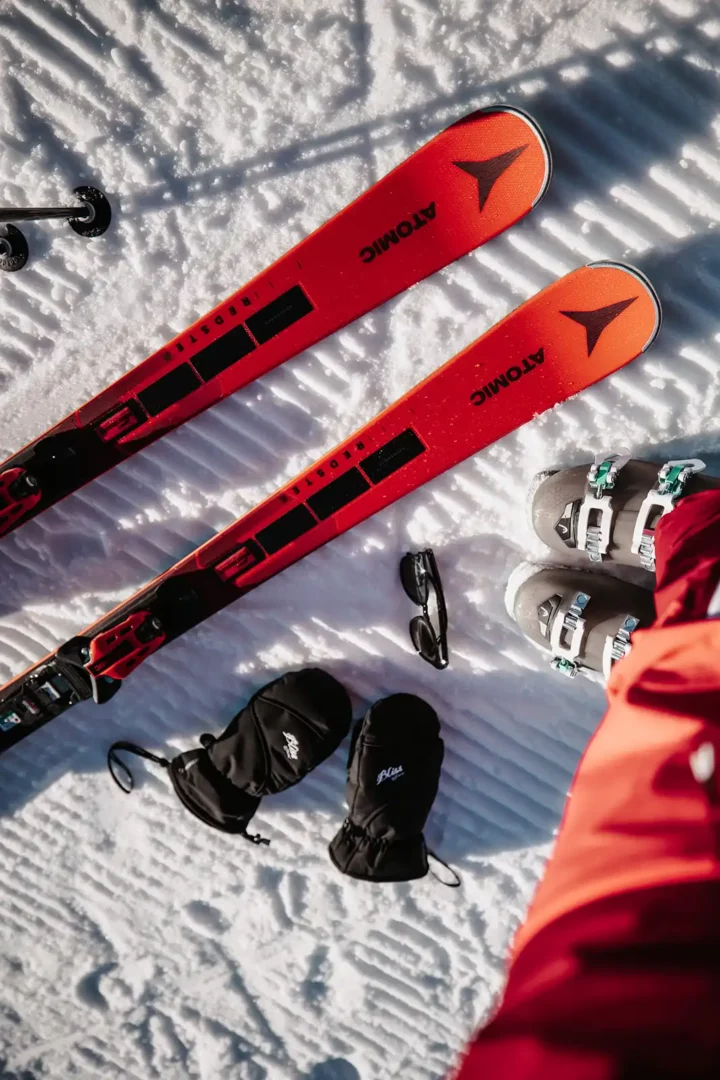 Skiequipment auf Schnee, roter Ski, Schwarze Handschuhe, schwarze Sonnenbrille, graue Skischuhe.