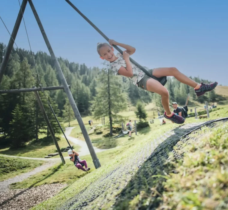 Dívka houpající se na zip line na dětském hřišti v Alpách, obklopená loukami, lesy a horami.