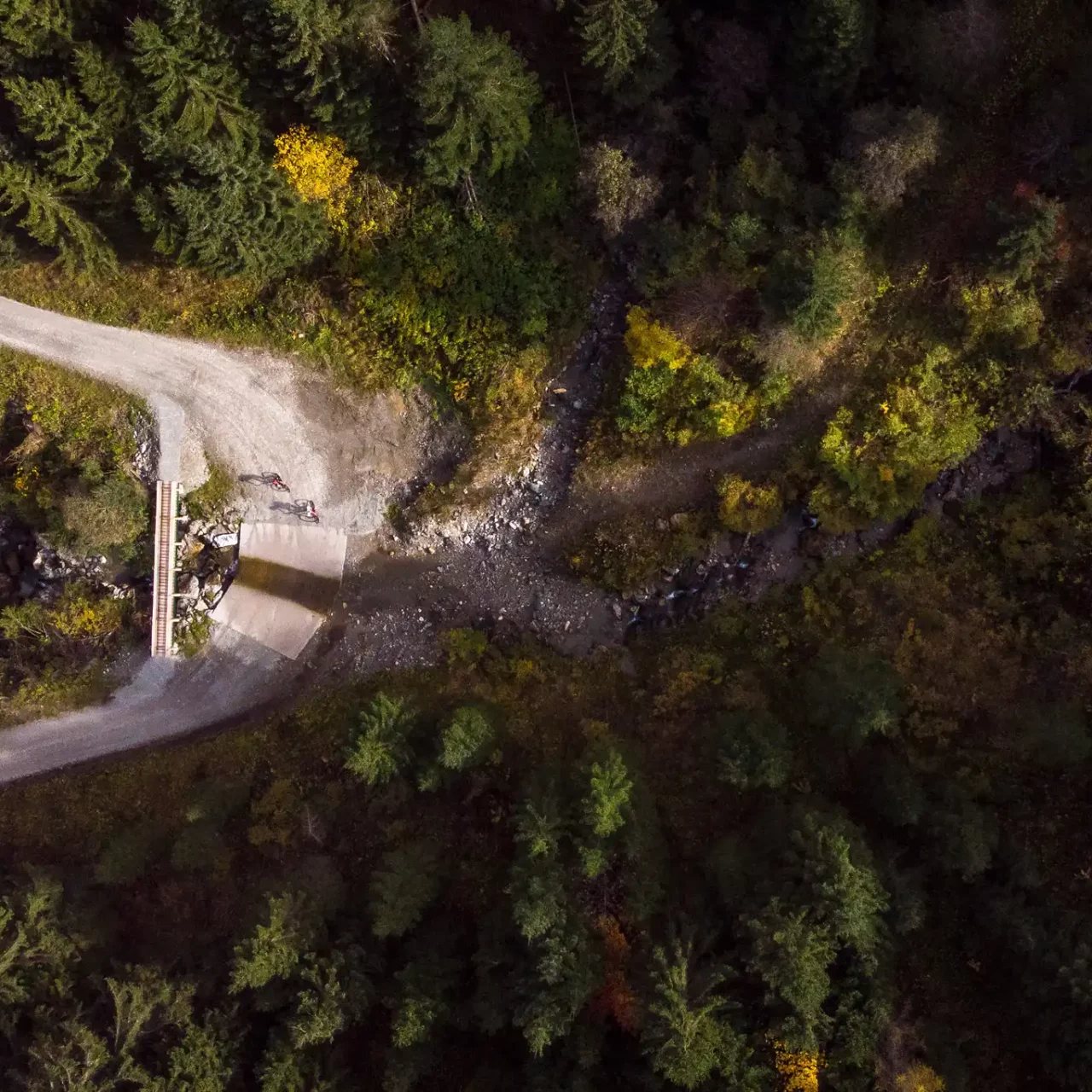 Záběr z dronu na dva horské cyklisty na lesní cestě s mostem přes horský potok v Salcbursku.