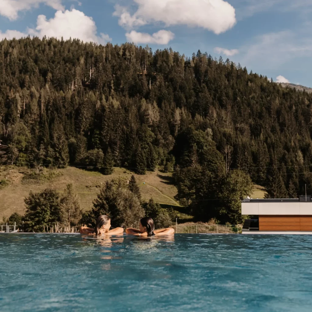 Hofgut-Wagrain-Apartment-Resort-Wagrain 615 Two guests enjoy the moment in the infinity pool at Hofgut Wagrain - with breathtaking views of the surrounding mountains and forests. Pure relaxation and alpine lifestyle combined in a very special place.