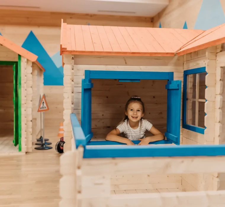 Spielerisches Entdecken im Mini Park: Ein fröhliches Mädchen blickt aus dem Fenster eines liebevoll gestalteten Spielhäuschens im Hofgut Wagrain – ein Ort voller Fantasie, Abenteuer und kindlicher Freude.