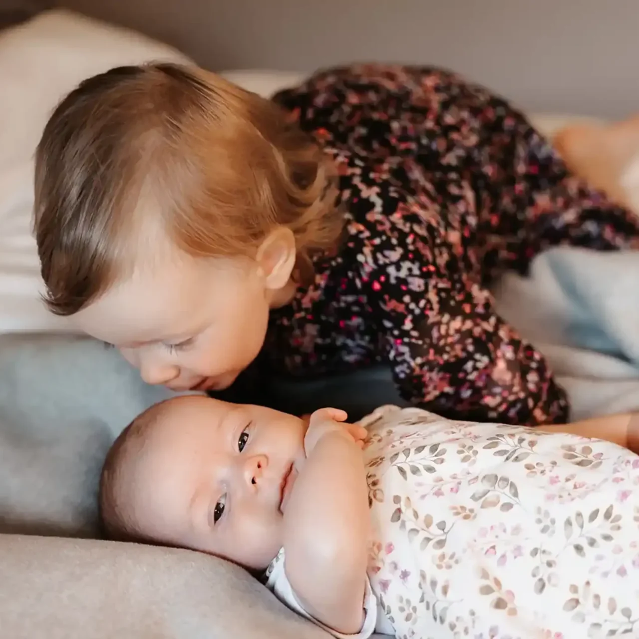Baby liegt wach auf einer grauen Decke, während ein Kleinkind in einem dunklen Blumenkleid im Hintergrund sitzt.