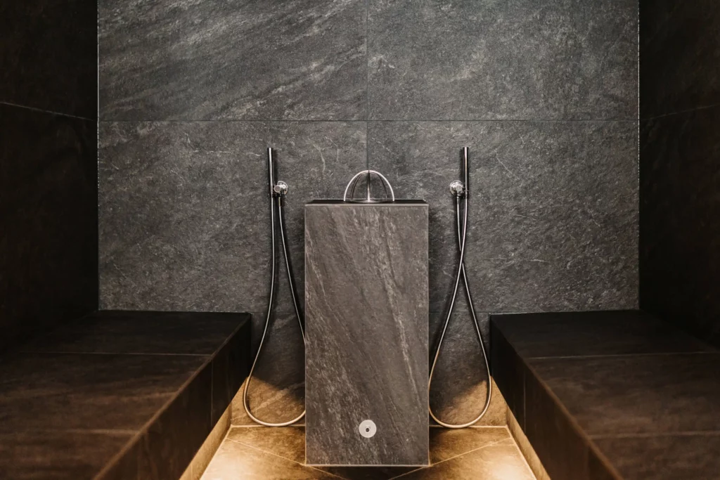 Hofgut-wagrain-apartment-adults-only-sauna-dampfbad-243 Moderner Dampfbadbereich mit dunklen Natursteinfliesen, zwei Duschköpfen und zentralem Wasserbecken für wohltuende Wellnessmomente.