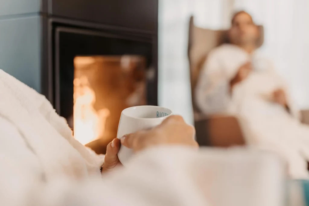 Hofgut-wagrain-apartment-adults-only-teepause-274 Nahaufnahme einer Tasse Tee in der Hand – im Hintergrund: flackerndes Kaminfeuer und entspannter Partner.