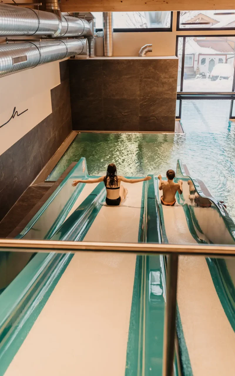 Hofgut-wagrain-apartment-sovielspaß-wasserrutsche-304 Zwei Kinder rutschen lachend nebeneinander auf der breiten TrioSlide ins warme Hallenbad des Hofgut Wagrain – Wasserspaß für kleine Gäste in sicherer, lichtdurchfluteter Umgebung.