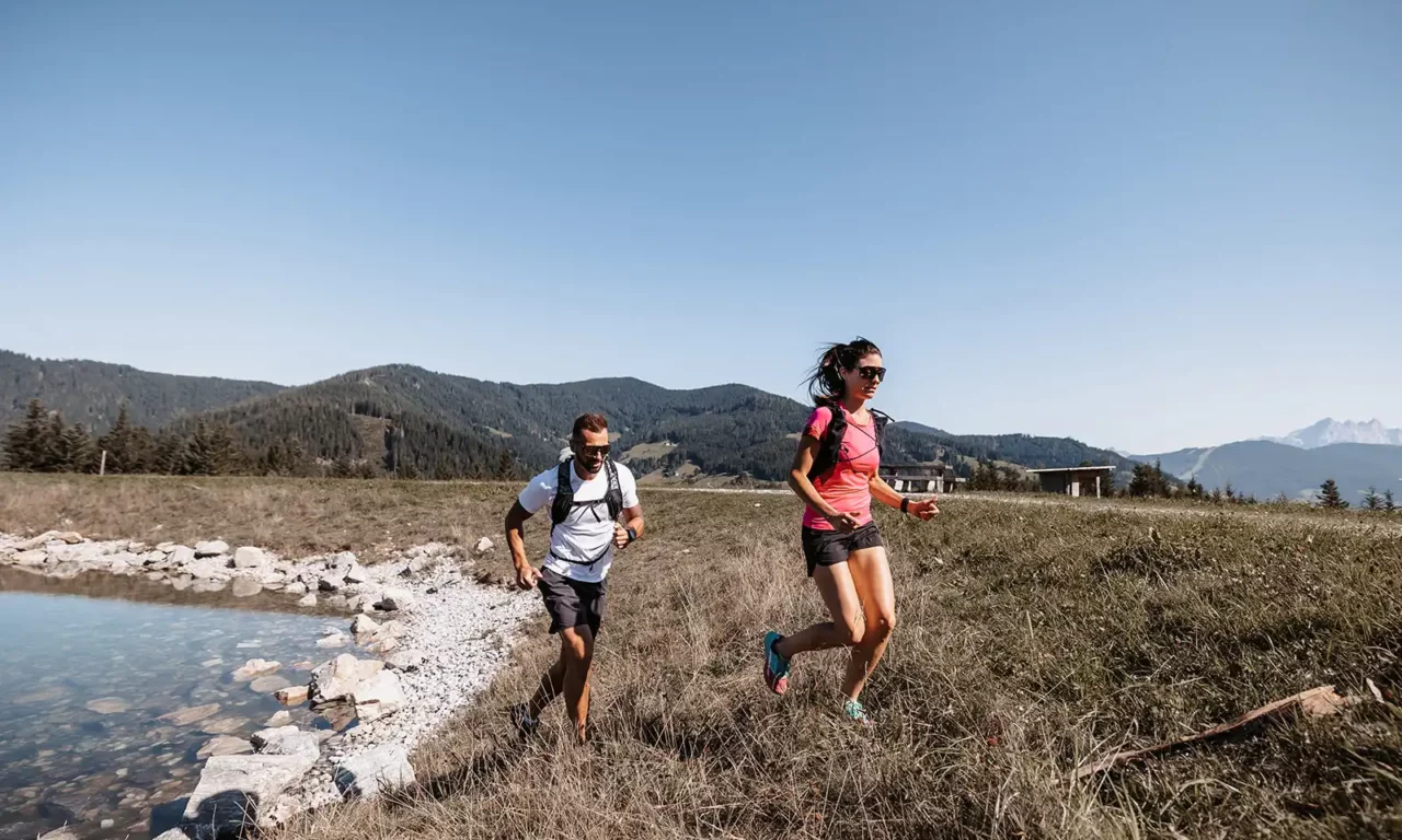 Hofgut©Elisabeth_Fotografie-253 An athletic couple with trail running backpacks jogs along a gravel path through the alpine landscape.