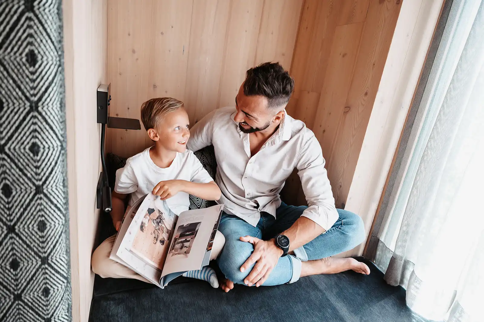 Hofgut©Elisabeth_Fotografie-386-Gutschein-schenken Father and son sit cosily in a wood-panelled alcove and leaf through a magazine together.