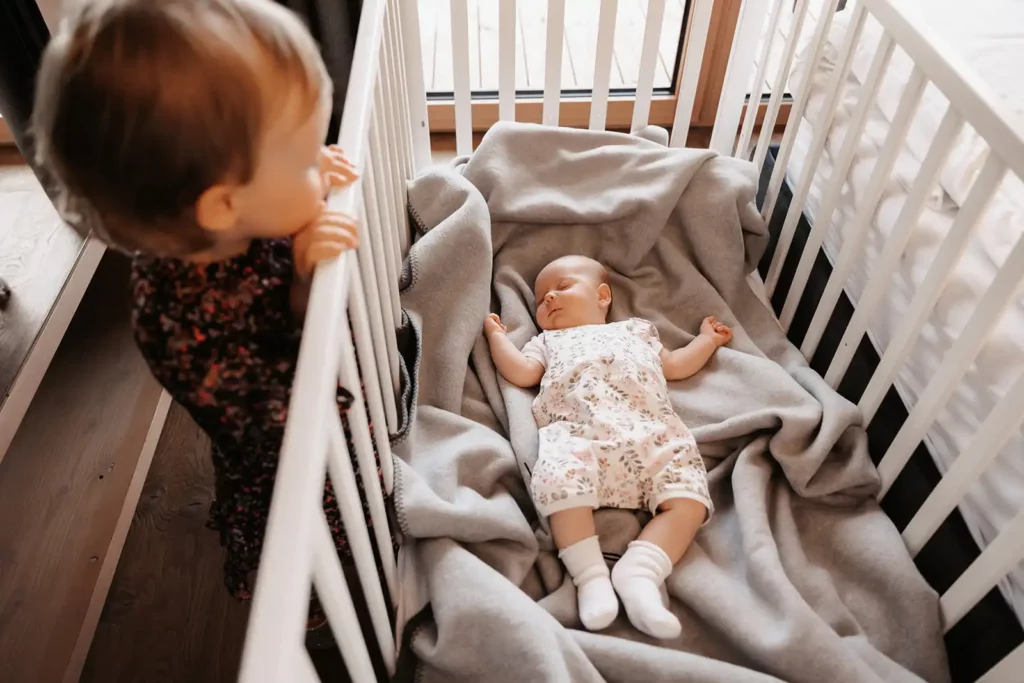 Hofgut©Elisabeth_Fotografie-471 A toddler looks curiously over the cot at a sleeping baby snuggled up in a grey blanket.