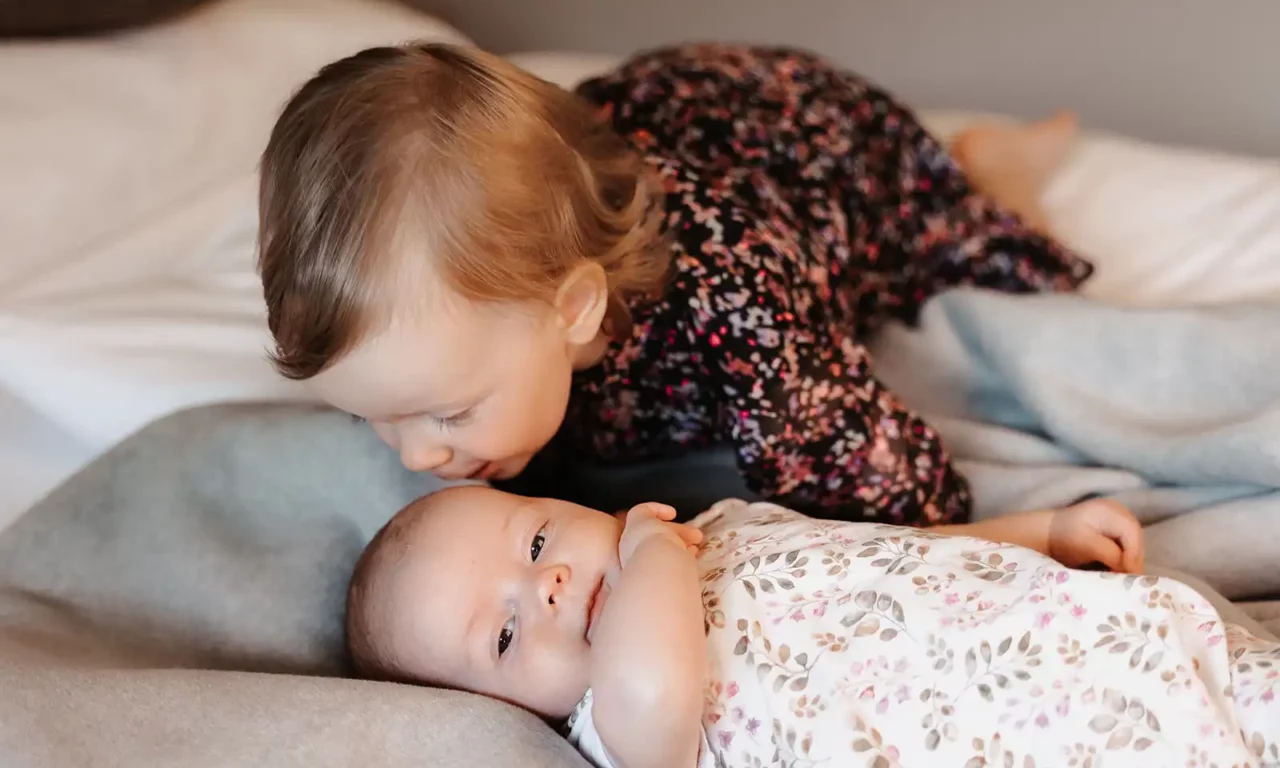 A baby is lying awake on a grey blanket while a toddler in a dark floral dress sits in the background.