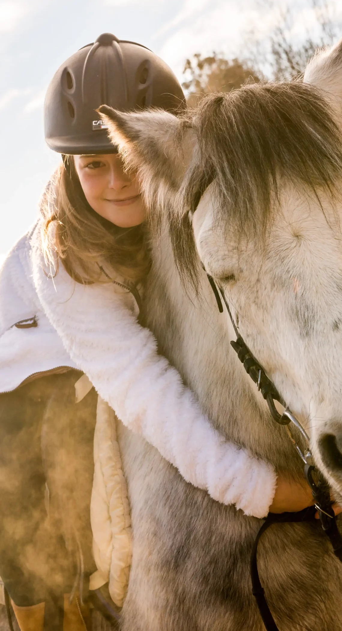 Ein Mädchen umarmt ein weißes Pferd auf einem Reitplatz, im Hintergrund ist ein Schild mit der Aufschrift "Postranch" zu sehen. Die Sonne scheint, und die Landschaft im Hintergrund ist von Bergen und Bäumen umgeben.