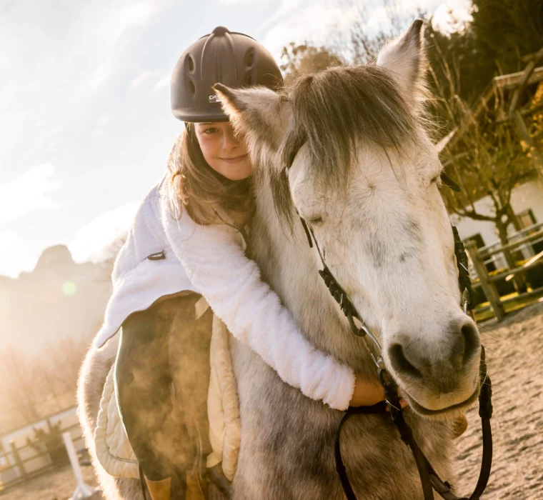 A girl is hugging a white horse in a riding arena, with a sign saying ‘Post Ranch’ in the background. The sun is shining and the landscape in the background is surrounded by mountains and trees.