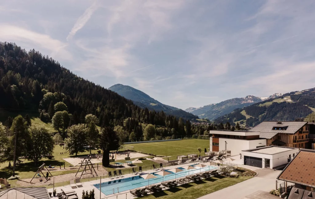 Weitläufige Außenanlage eines Resorts mit Outdoor-Pool, Liegebereich, Spielplatz und großzügiger Grünfläche – eingebettet in die Berglandschaft des Salzburger Landes mit Ausblick auf bewaldete Hänge und Gipfel.