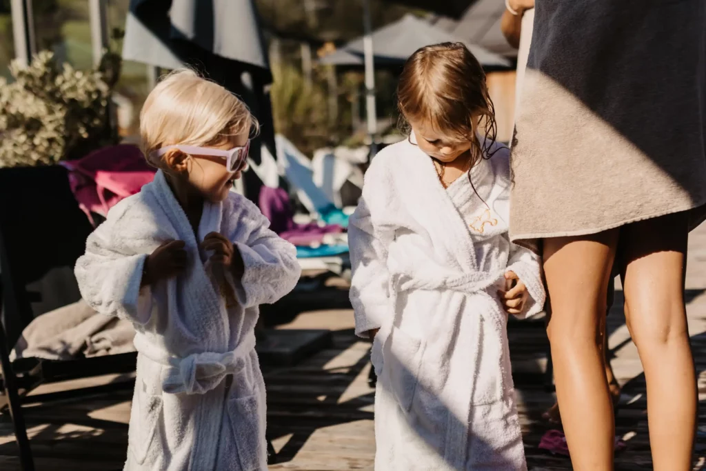 Two small children in white bathrobes stand next to each other on a sun terrace by the pool - one is wearing sunglasses, the other has wet hair, summer relaxation at the family resort.