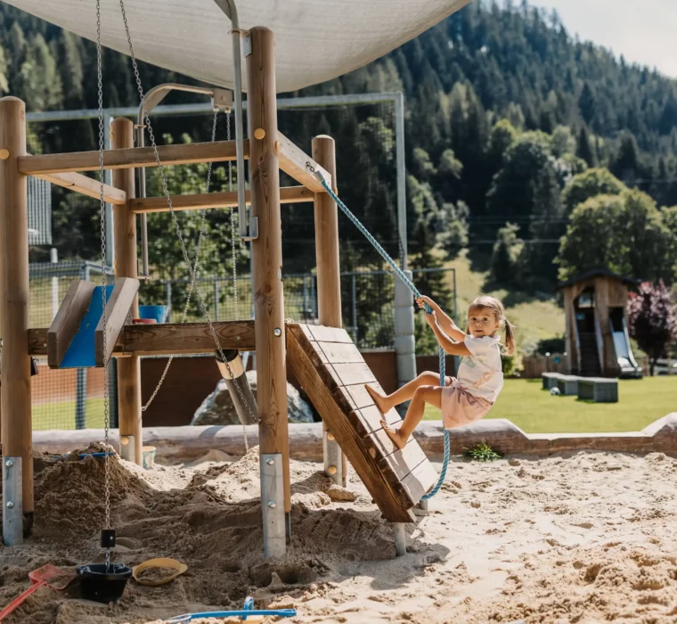 Hofgut-Wagrain-Apartment-Resort-Wagrain-spielplatz-sankiste- 484 Spielspaß pur im Hofgutgarten des Hofgut Wagrain – ein kleines Mädchen entdeckt barfuß die Kletterlandschaft im Sandspielplatz vor traumhafter Alpenkulisse.