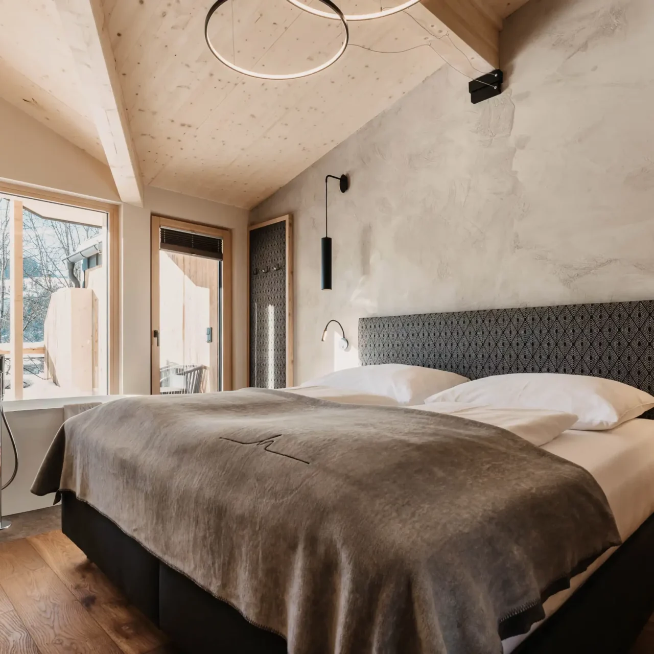 Modern eingerichtetes Schlafzimmer mit gemütlichem Doppelbett, freistehender Badewanne vor bodentiefem Fenster und stilvollem Holzdach – alpiner Luxus mit Panoramablick.