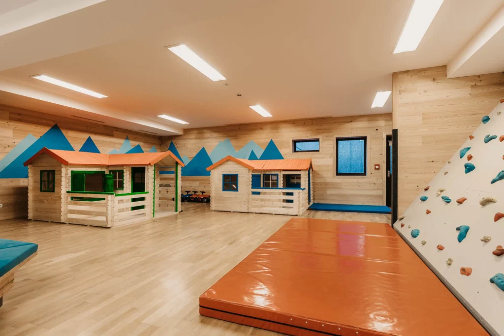 Moderner Indoor-Spielbereich mit kleinen Holzspielhäusern, Kletterwand und weicher Matte – kindgerechte Ausstattung in heller, freundlicher Umgebung mit Bergmotiv an der Wand.