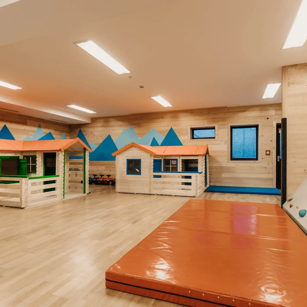 Moderner Indoor-Spielbereich mit kleinen Holzspielhäusern, Kletterwand und weicher Matte – kindgerechte Ausstattung in heller, freundlicher Umgebung mit Bergmotiv an der Wand.