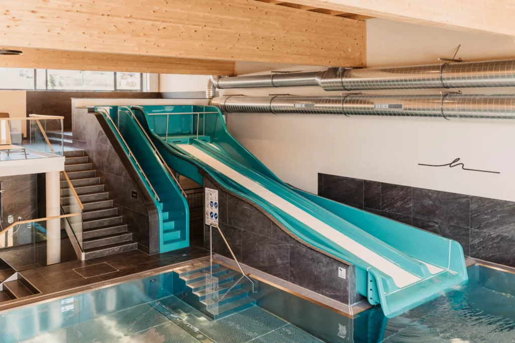 Indoor family pool area with large blue water slide and modern equipment - wide slide channel ends in the heated pool, surrounded by stylish design and wooden ceiling.