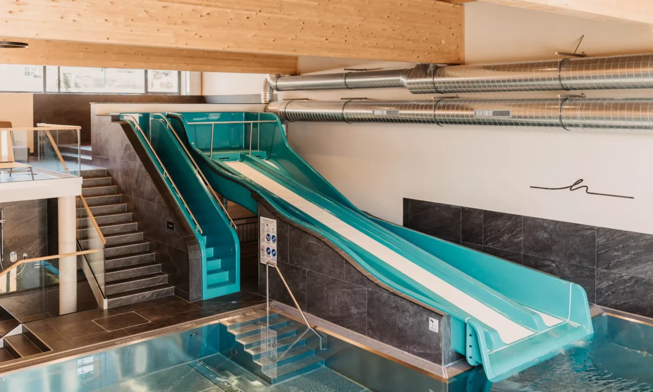 Innenbereich eines Familienpools mit großer blauer Wasserrutsche und moderner Ausstattung – breiter Rutschkanal endet im beheizten Becken, umgeben von stilvollem Design und Holzdecke.