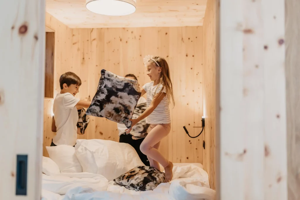 Kinder spielen fröhlich auf einem Bett in einem gemütlichen Zimmer mit Holzverkleidung – Kissen fliegen durch die Luft bei einer ausgelassenen Kissenschlacht.