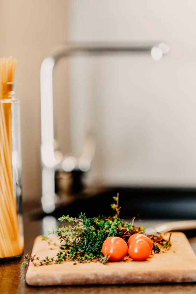 Hofgut_Apartement1-35 Frische Cocktailtomaten und Kräuter auf Holzbrett in moderner Apartmentküche mit Pasta im Hintergrund