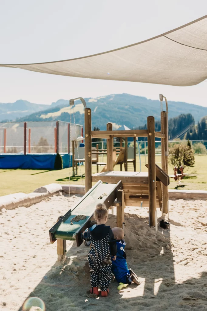 Hofgut_Spielplatz Two small children playing in the sand area of an outdoor playground with wooden scaffolding and water channel - shady area with a view of green meadows and mountains in the background.